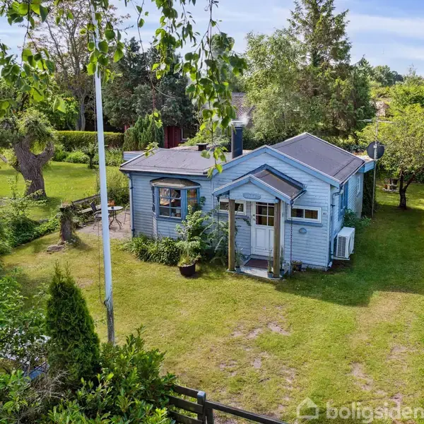Et lille, blåt hus med veranda står i en frodig have omkranset af buske og træer. En flagstang står foran huset, der har en grussti og grønne omgivelser.