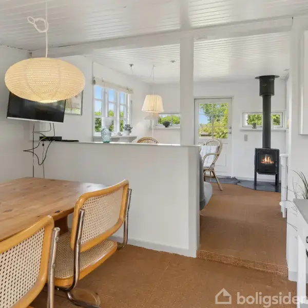 Træbord omgivet af flettede stole i en spisestue med lamineret hvide vægge. En loftslampe, et fjernsyn på væggen, og en brændeovn ses i baggrunden.