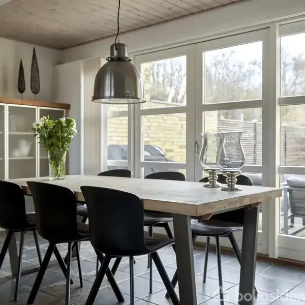 Et træbord med sorte stole står i et lyst køkken/alrum. En blomsterbuket er placeret på bordet, mens glasvaser står ved siden af. Store vinduer viser en terrasse udenfor.