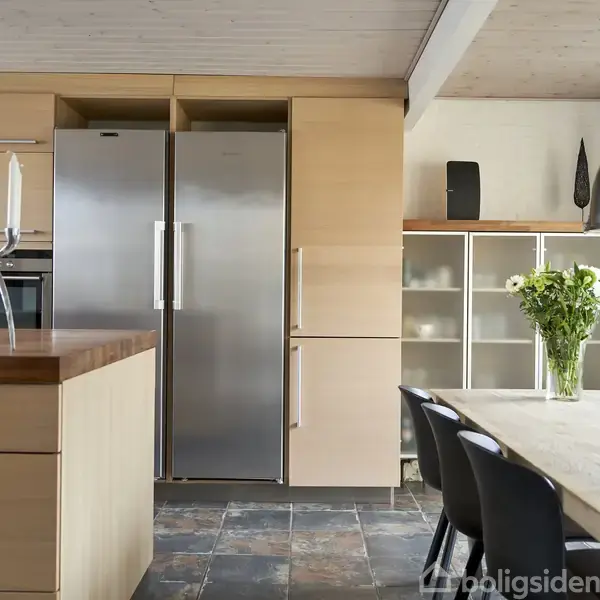 Køkken med moderne træskabe, to stålkøleskabe, en stor lys spisebord med sorte stole. En vase med friske blomster står på bordet, omgivet af minimalistiske dekorationer.