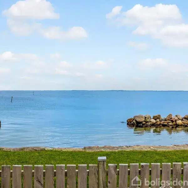 Et træhegn står stille foran en rolig strand med en stenbrud mod en blå himmel og hav i baggrunden.