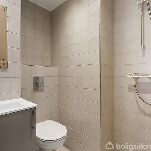 Badeværelse med lyse fliser, en bruser på væggen samt en håndvask til venstre. Toilettet er indbygget, og der er en spejlkant i venstre side.