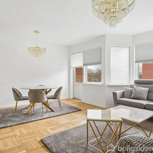 En moderne stue med en grå sofa og sofabord, en spisebord med fire stole. Parketgulv og to store vinduer med gardiner. En lysekrone hænger fra loftet.