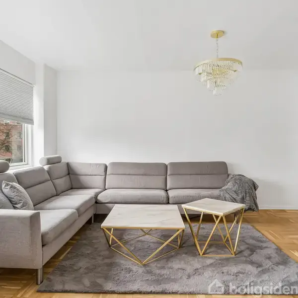 Grå hjørnesofa står i stue med parketgulv, lysindfald fra vindue med persienner, gyldent sofabord over gråt tæppe. Hvid væg og loft med lysekrone i minimalistisk indretning.