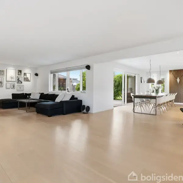 Stue med sorte sofaer og sofabord, åbner op til spisestue med hvidt bord og stole. Store vinduer giver udsigt til en have. Vægmonterede billeder dekorerer væggen.