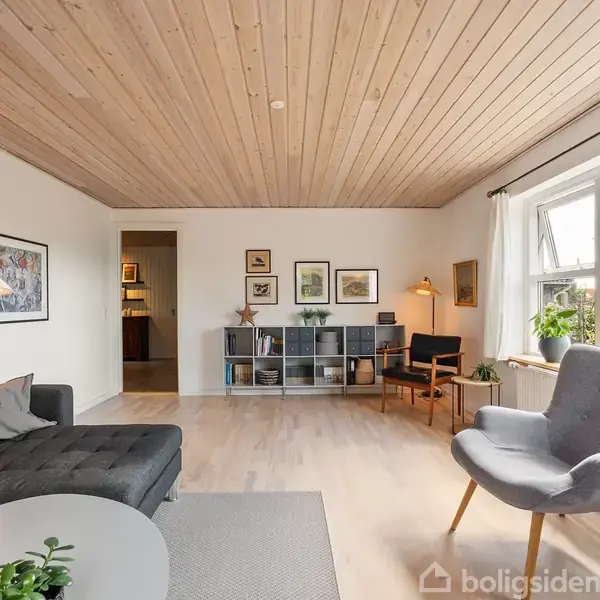 En stue med moderne møblering, herunder en grå sofa, lænestol, og reol. Væggene er dekoreret med billedrammer. Store vinduer lader lys komme ind i det lyse, åbne rum.