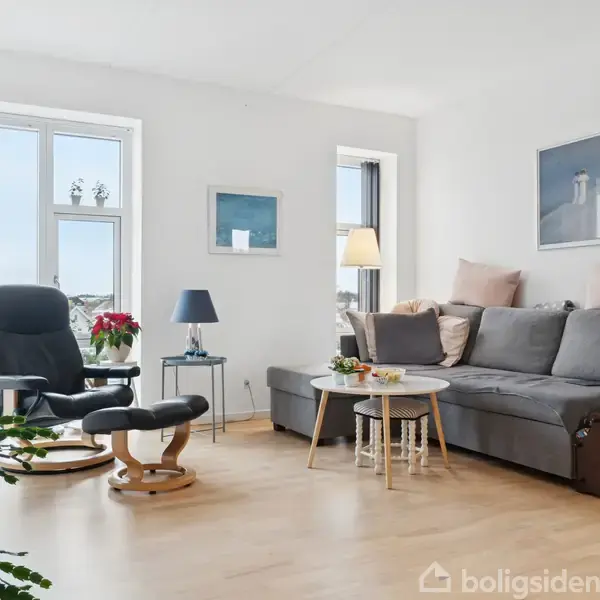 Gråt sofasæt med puder står i en lys stue. Ved siden af er et rundt sofabord med en lampe og dekoration. Store vinduer giver udsigt. Der er planter og billeder på væggene.