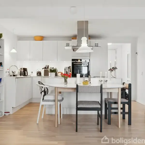 Køkken med et rundt træbord, omgivet af forskellige stole. Bordet er dækket af blomster og lysestager. I baggrunden er der hvide skabe og moderne apparater i et lyst rum.