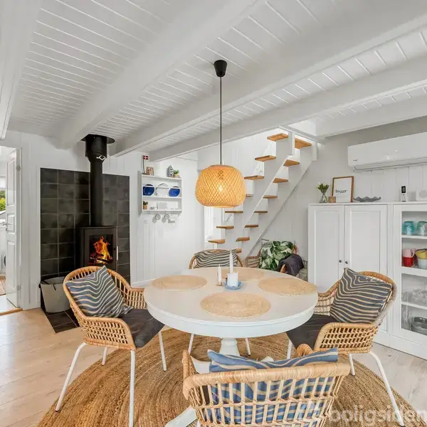 Et lyst rum indeholder et rundt spisebord med flettede stole omkring, ved siden af en tændt brændeovn. Trappen fører til en overetage, mens et vitrineskab viser porcelæn.