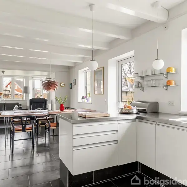 Et moderne køkken med hvide skabe og sorte fliser, bordplader er stilfuldt indrettet. Spisebordet bagved er pyntet med stole og en lampe. Store vinduer giver rigeligt naturligt lys.