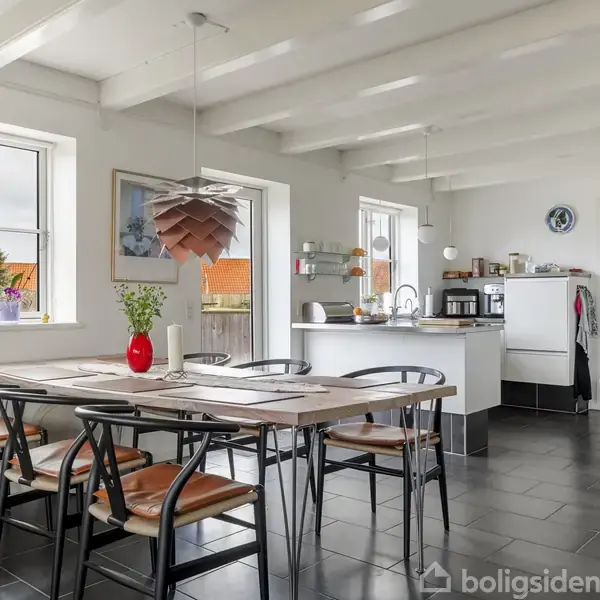 Et spisebord omgivet af sorte stole står opstillet i et lyst køkken-alrum. Rummet har hvide vægge, mørkt gulv, og der hænger en moderne lampe over bordet.