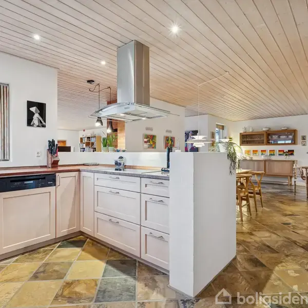 Et lyst køkken med træskuffer og integreret emhætte. Rummet er åbent med flisegulv, og der er spiseplads og dekorative elementer som lamper og planter i baggrunden.