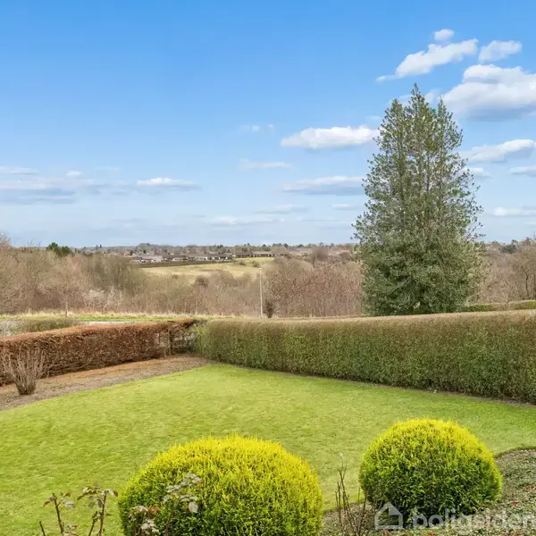 En velplejet have med grønne buske og en stor græsplæne; omgivet af hække. I baggrunden ses en enkelt høj træ og en åben landlig udsigt under en klar blå himmel.