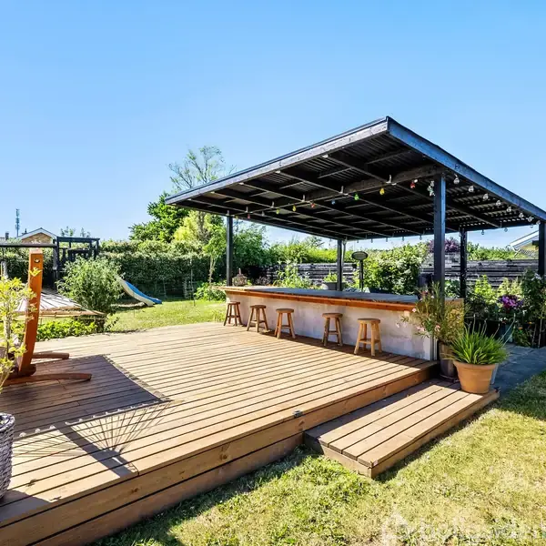 Terrasse med trægulv rummer barområde med fire barstole under et overdækket tag. Planter omkranser terrassen, og der er græs på plænen omkring. Himlen er klar og blå.