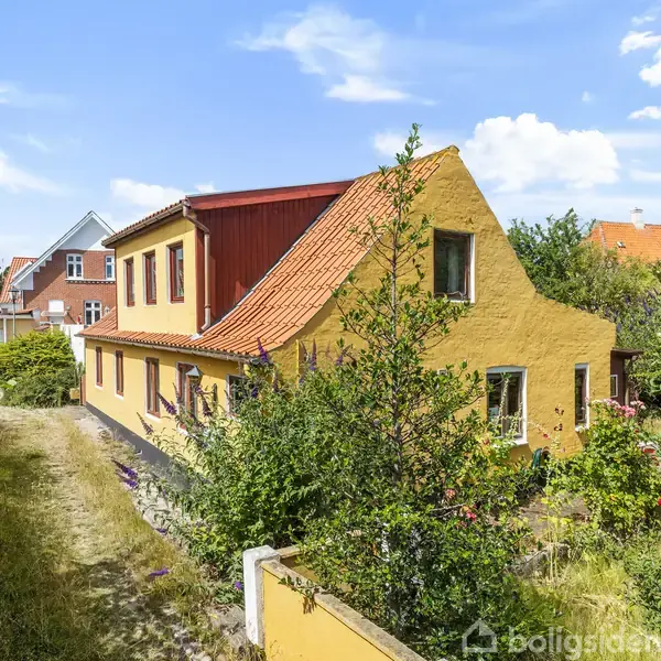 Gulmalet hus med rødt tegltag, omgivet af frodige buske og træer. Det ligger i et boligområde med andre lignende huse i baggrunden under en blå himmel med få skyer.