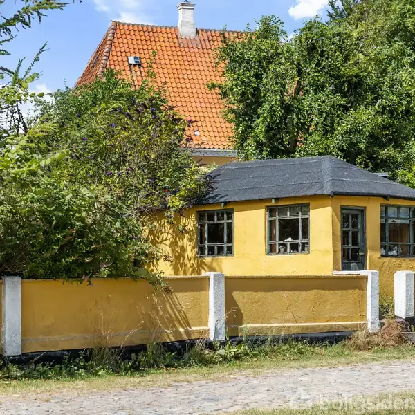 Gult hus med sort tag står stille i grønt landskab omgivet af tætte buske og træer. Rødtagede bygninger ses i baggrunden under en blå himmel.