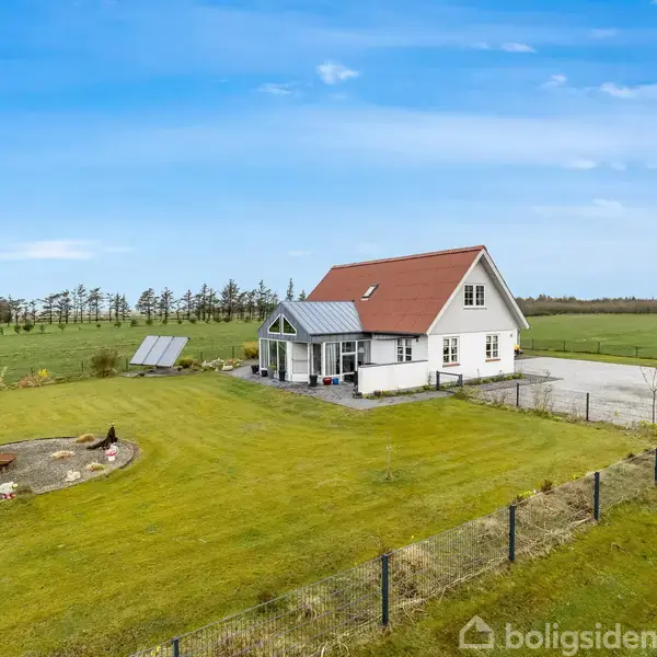 Et hvidt hus med rødt tag står på en stor græsplæne med en indhegning. To solpaneler er placeret tæt på huset. Træer ses i baggrunden under en blå himmel.