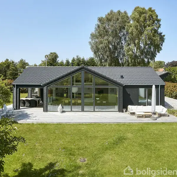 En moderne, sort ensplansvilla med store vinduer står stille på en solrig træterrasse i en grøn have omkranset af buske og træer.
