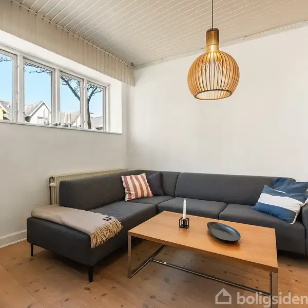 En grå sofa står i et hjørne med puder og et tæppe. Foran er et træbord med en lysestage og skål. En lampe hænger over bordet. Vinduer giver naturligt lys.