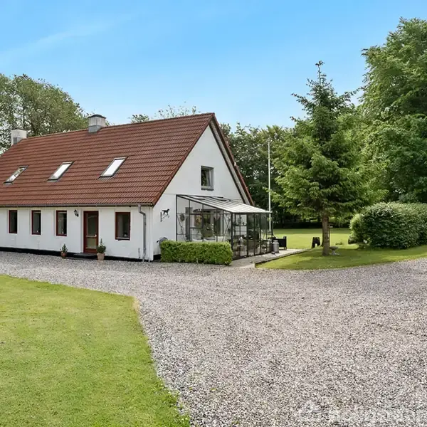 Hus med rødt tag står ved en grusvej omgivet af græsplæner og store træer. En terrasse er synlig ved siden af huset. Skyfri blå himmel ovenover.