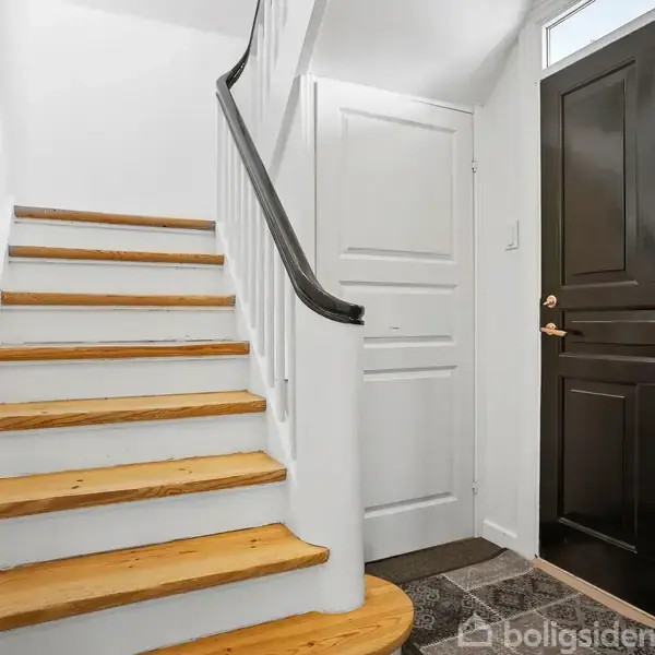 Trætrappe fører opad med sorte gelændere; sort hoveddør med guldbeslag ved siden af, i en gang med lyse vægge og flisegulv.