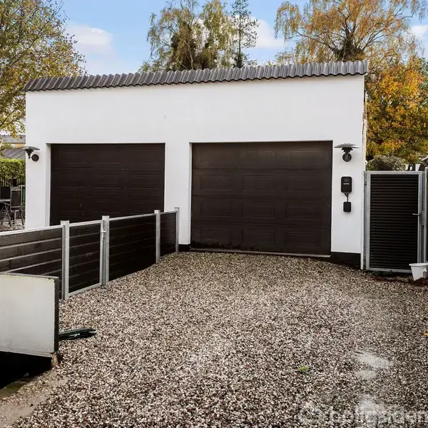 To garagedøre i en hvid bygning står lukkede, omgivet af grusbelagt indkørsel og mørke hegner; træer og nabohus ses i baggrunden.