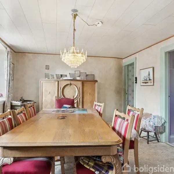 Langbord med stole står i en spisestue. En lysekrone hænger over bordet. Rummet har et skab i baggrunden og lyse vægge. Vindue med gardiner til venstre. Ingen tekst til stede.