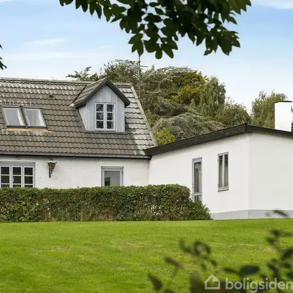Et hvidt hus med skråt tag står ved siden af en carport med en bil. Huset er omgivet af en hæk og grønne træer under en blå himmel.
