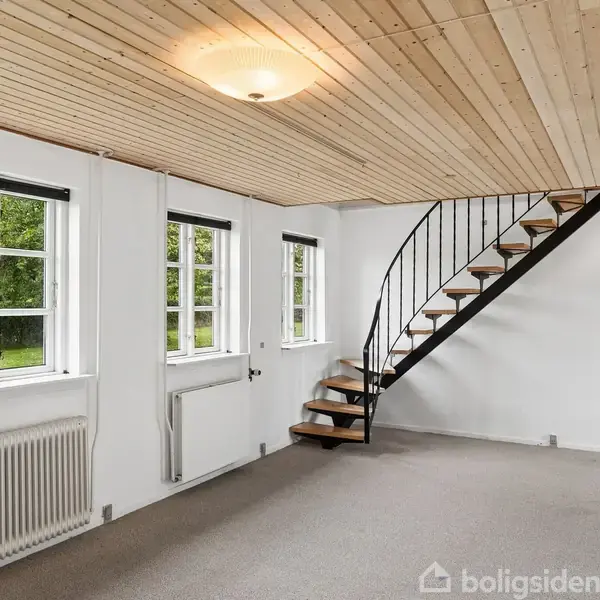 Et rum med en trappe, der fører opad. Træloftet har en enkel lampe. Tre vinduer med udsigt til grønne omgivelser og en radiator under vinduerne. Væggene er hvide.