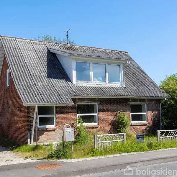 Et rødt murstenshus med et gråt, skråt tag står i en frodig have ved en stille vej. Foran huset er der en lille græsplæne og en indkørsel.