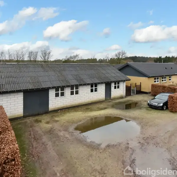 Bygning med gråt tag og hvide mure står i en gårdsplads med grus, omgivet af hæk. Ved siden af er en bil parkeret. I baggrunden ses flere huse og træer.
