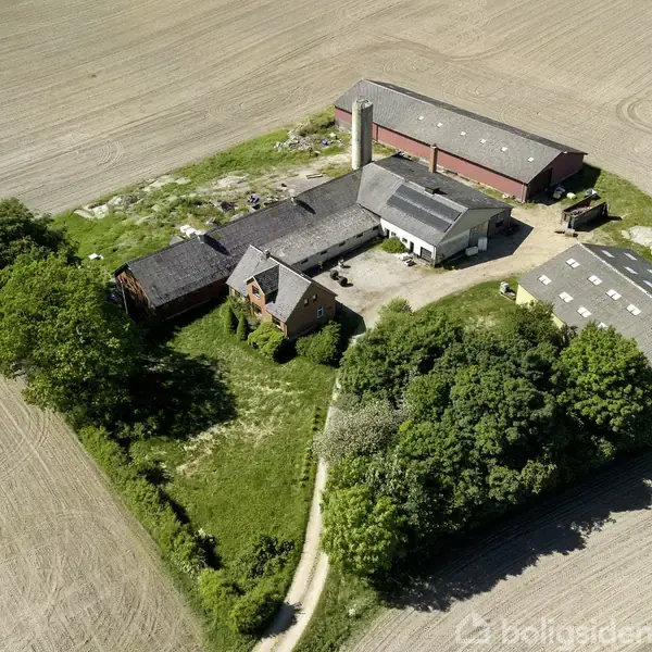 Gårdkompleks med flere bygninger, herunder en lade og silo, omgivet af marker og en grusvej. Træer og buske gror tæt ved bygningerne, der danner en firkantet gårdsplads.