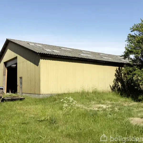 En gul lade står stille på en græsmark med træer i baggrunden under en klar blå himmel. Der er ingen tekst i billedet.