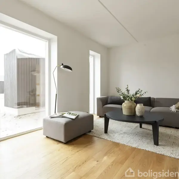En stue har en grå sofa med puder og et rundt, sort sofabord med planter. Et stort vindue og en dør lukker lys ind fra en terrasse. Gulvet er af træ.