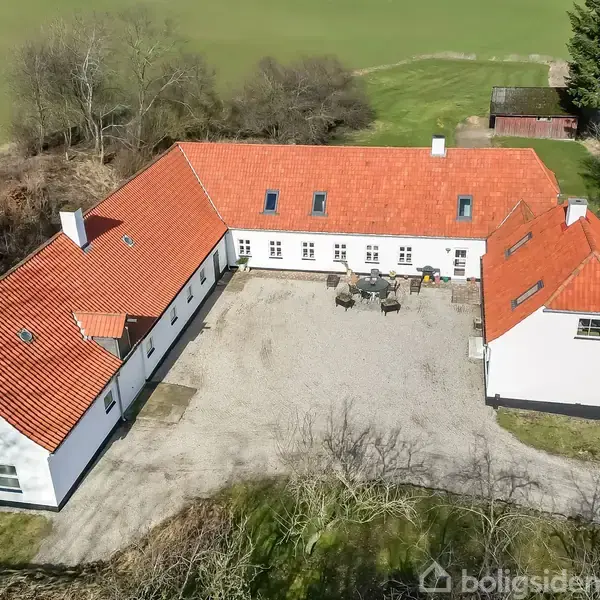 En L-formet gårdbygning med hvidt murværk og rødt tegltag omkranser en gårdsplads med havemøbler. Omgivet af marker og træer, ligger ejendommen i landlige omgivelser.