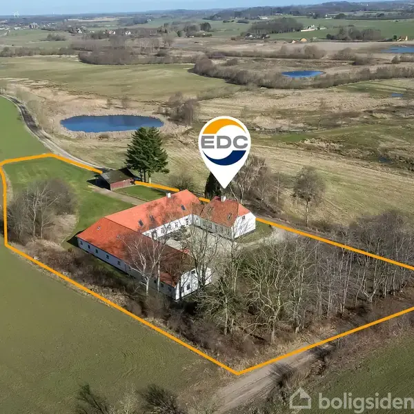 Bygning med rødt tag markeret med "EDC," omgivet af marker og træer. En orange linje afgrænser ejendommen, som ligger i et åbent, landligt område med små søer i baggrunden.