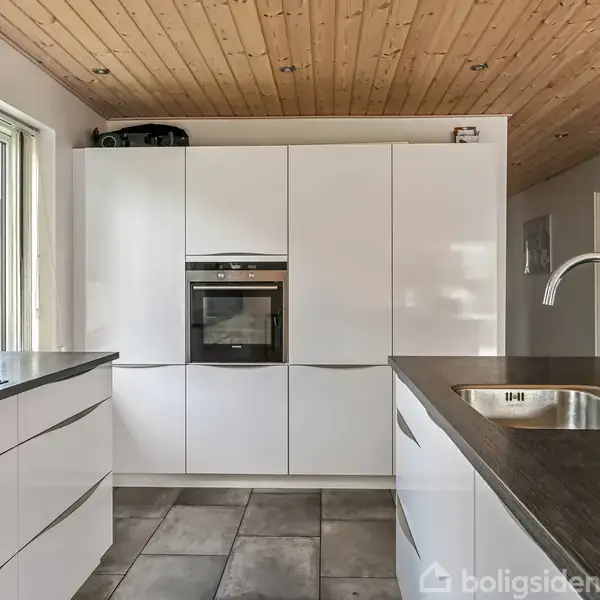 Et moderne køkken med hvide skabe og en indbygget ovn. En skinnende vandhane og vask er monteret i den mørke bordplade. Træloft og flisegulv skaber en lys, stilfuld atmosfære.