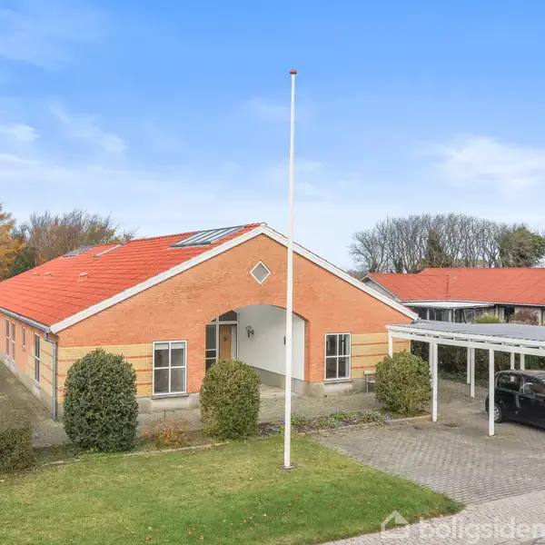 Rødstenshus med rødt tag står på en velplejet græsplæne. Et hvidt flagstang står foran huset, og en carport med en parkeret bil ligger ved siden af. Træer ses bagved.