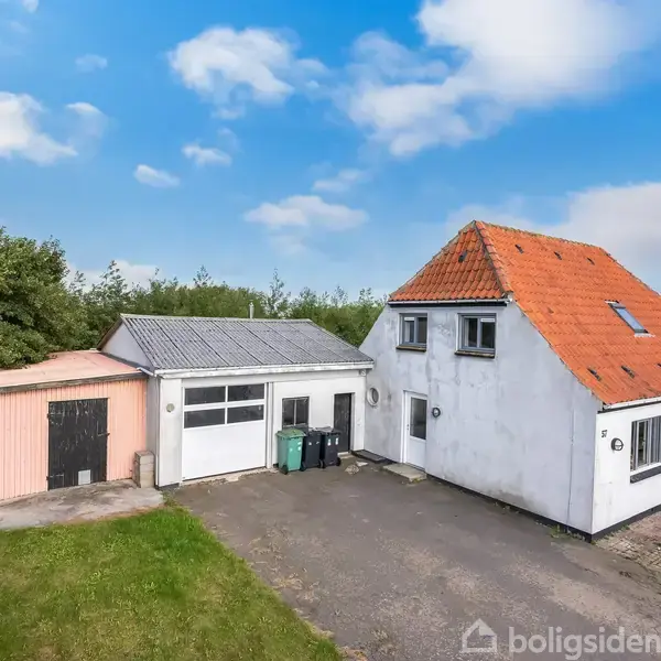 Et hvidt hus med rødt tag står i en indkørsel ved siden af en garage og et lille udhus. Græs og buske omgiver bygningerne under en blå himmel.