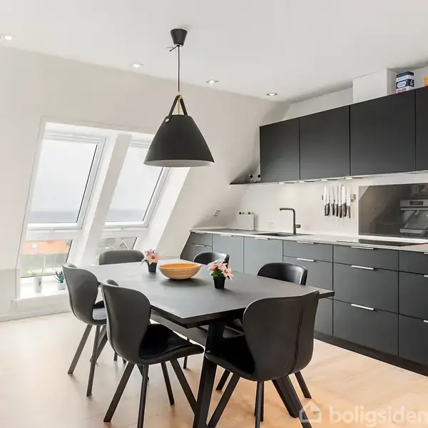 En stue med sofa og lænestol arrangeret på et tæppe, foran et tv. Rummet har skråvinduer og åbner mod en balkon samt et køkken i baggrunden.