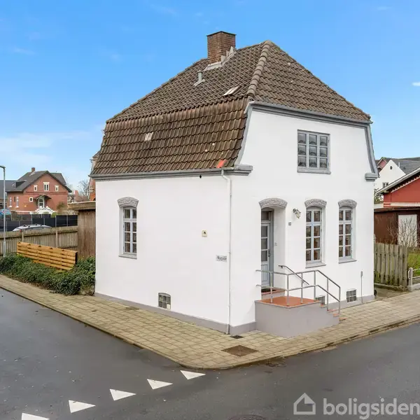 Hvidt hus med mørkt tegltag står tomt på et gadehjørne. Huset har trapper ved hoveddøren, små vinduer og er omgivet af et lille hegn. Veje og andre huse ses omkring.