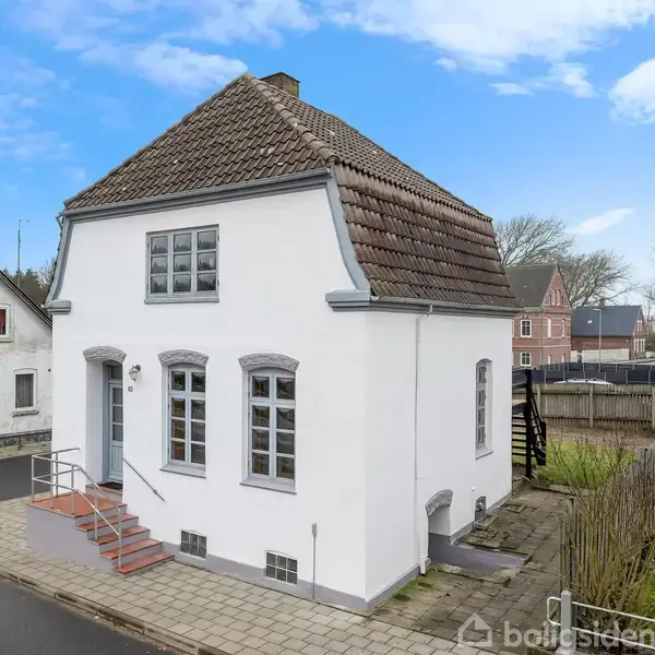 Hvidt hus med brun tegltag placeret på en hjørnegrund. Huset har flere vinduer og en lille trappe ved hoveddøren. Omgivet af fortov, græs og nærliggende bygninger.