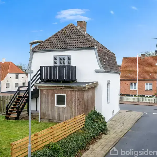 Et hvidt hus med en træterrasse og udendørs trappe står i en rolig byområde. Baggrund med røde taghuse og en traditionel mølle. Græsplæne omkranset af et hegn.