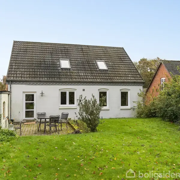 Et hvidt hus med mørkt tag og to tagvinduer står i en grøn have. En terrasse med havemøbler er placeret foran huset, omgivet af buske og træer.