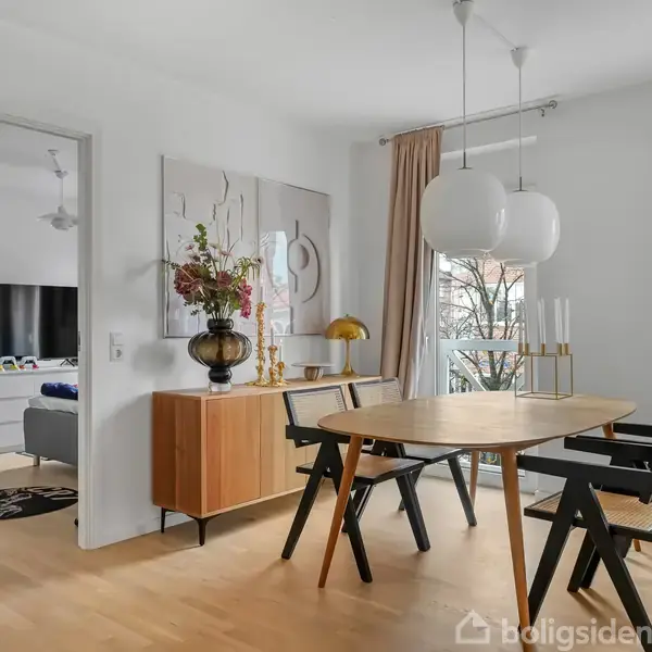 Et spisebord med fire stole står i et lyst rum. En kommode med blomster står ved væggen. En åben dør fører til en stue med en sofa og fjernsyn.