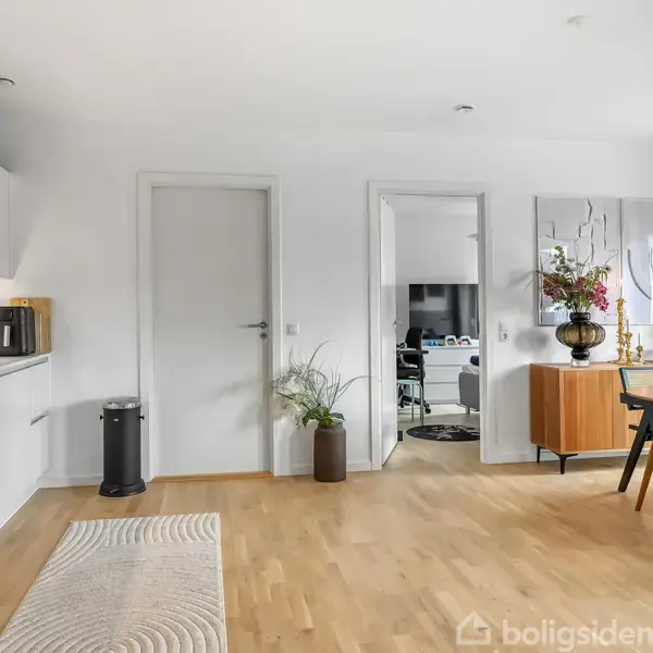 Køkken-alrum med lyse trægulve, to døre, spisebord med stole, køkkenskabe til venstre og en kommode med vase til højre. Store loftslamper hænger over bordet.