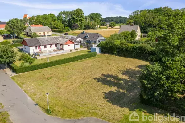 En græsbeklædt grund foran et hvidt hus med rødt tag, omgivet af hække og flere huse i baggrunden, i en landsbyagtig setting med frodig natur.