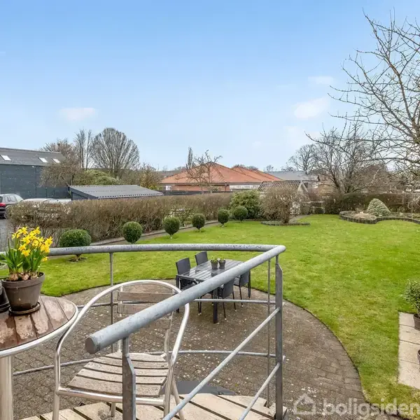 Bord med potteplanter står på en terrasse, omgivet af grøn have med hække. Der er havemøbler på græsplænen, et drivhus til højre, og parkerede biler i baggrunden.