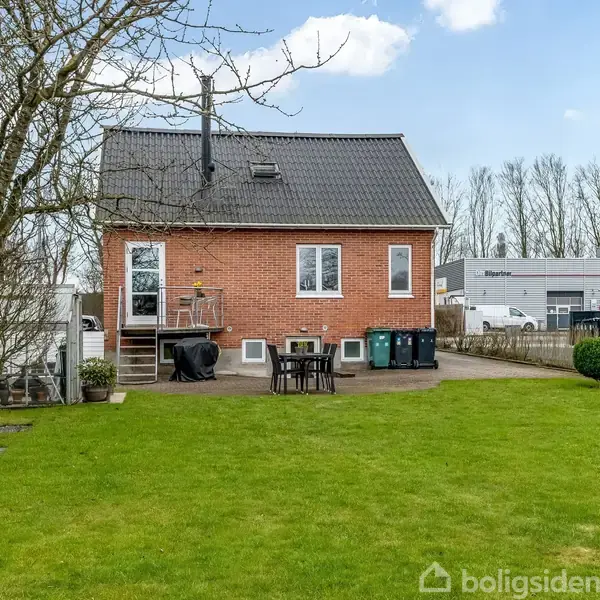 Rødt murstenshus med trappe og terrasse, står i en velholdt have. Omkring huset er der græsplæne, træer, og hække. Bagved ses flere bygninger og en klar himmel.