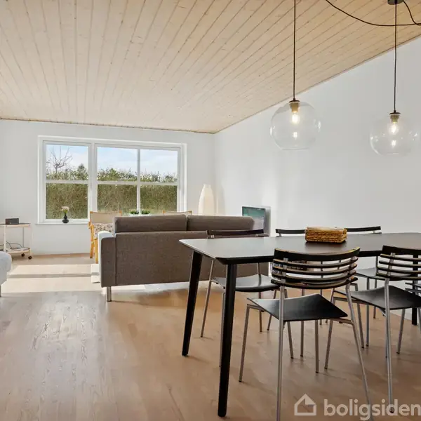 Moderne stue med sofagruppe ved stort vindue, der giver udsigt til have. Spisebord med stole står under to hængelamper. Lys træloft og gulv skaber en hyggelig atmosfære.